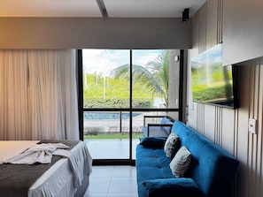 A modern hotel room with a bed, a blue sofa, a TV mounted on the wall, and a large sliding glass door leading to an outdoor area. - Makia Beach Experience Muro Alto (Ipojuca)