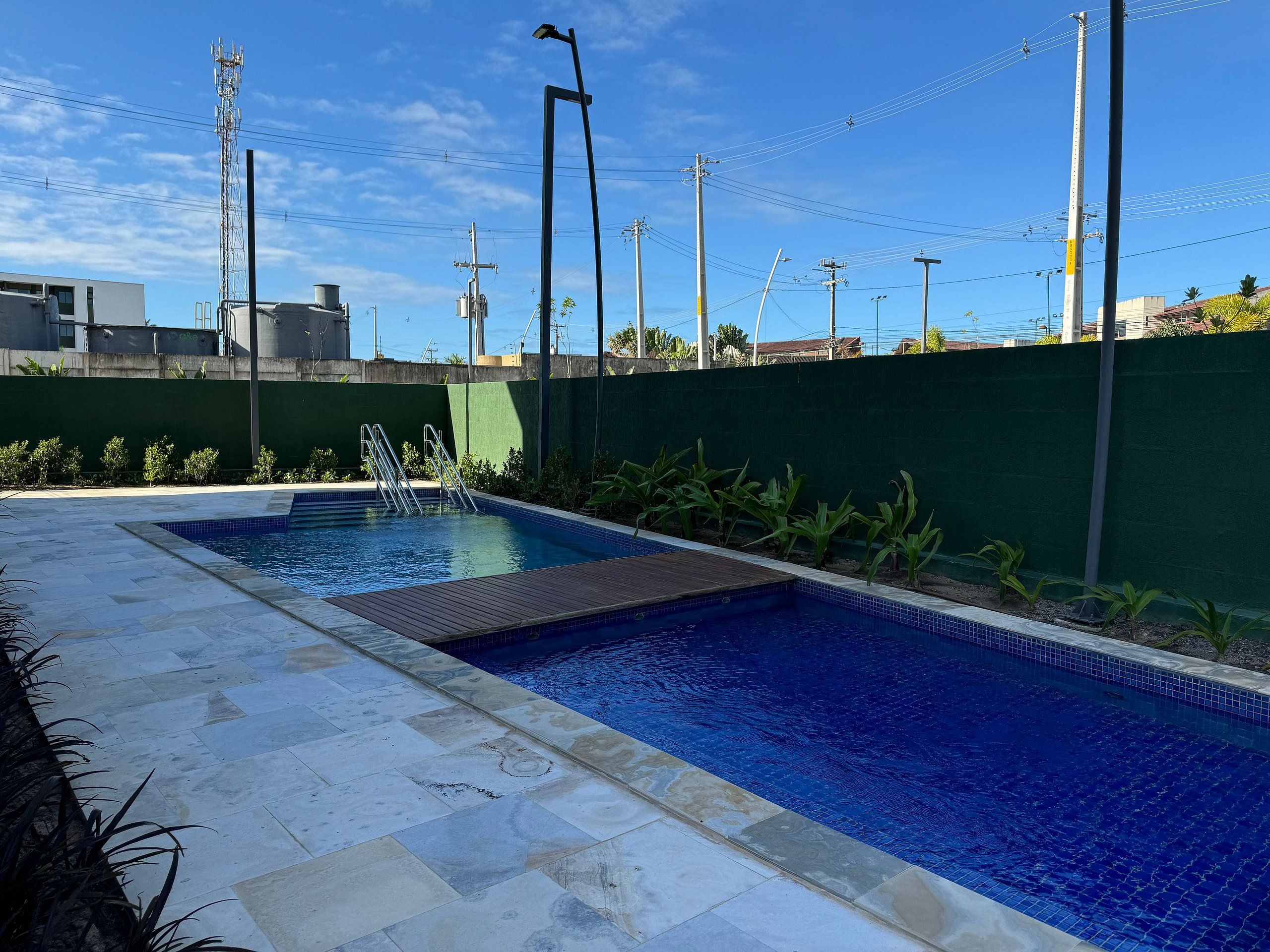 Una piscina con solárium empedrado, rodeada por un muro verde y plantas.