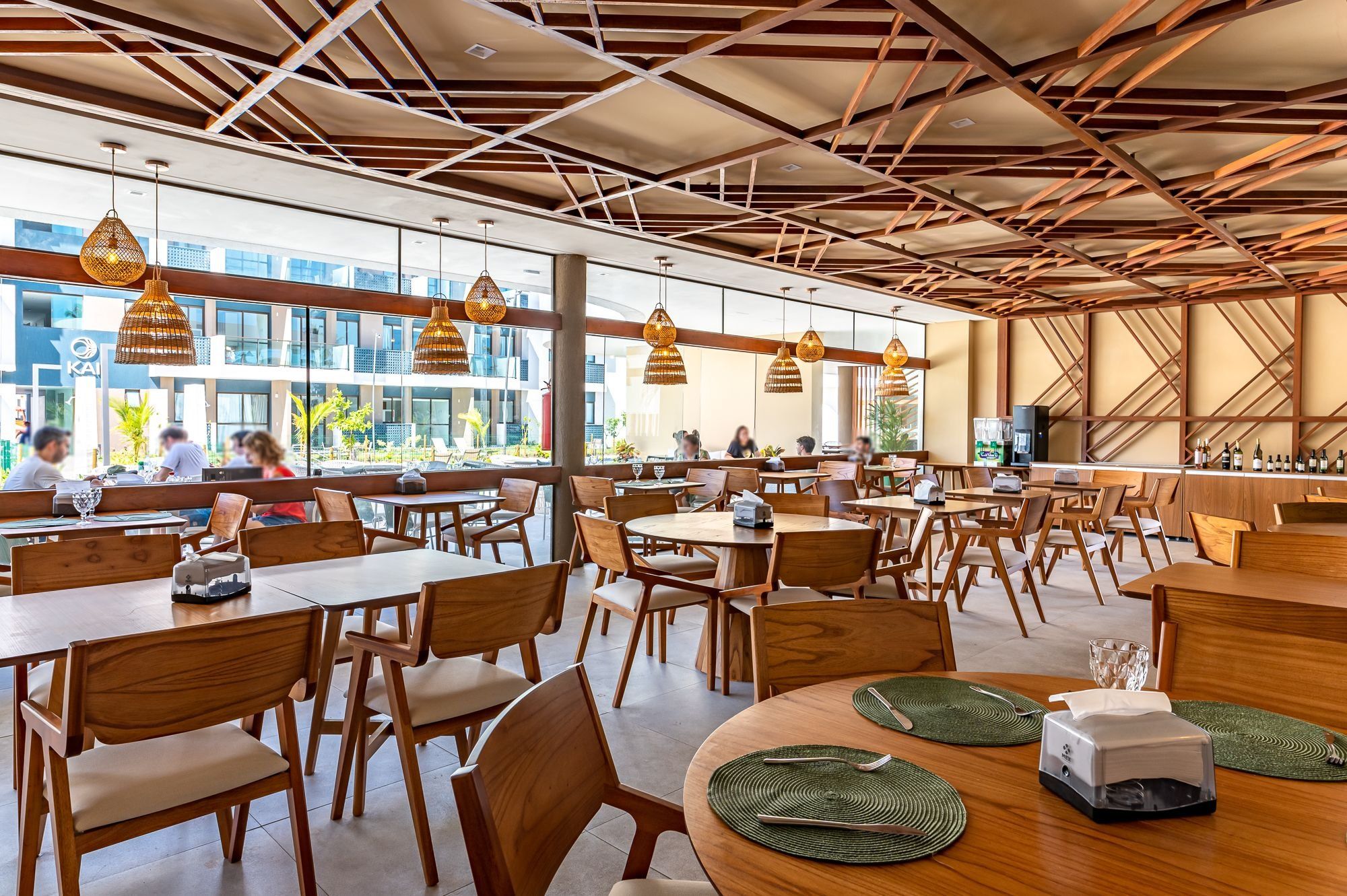 Un amplio comedor interior con varias mesas y sillas, un diseño moderno en el techo y grandes ventanales que ofrecen vistas al exterior.