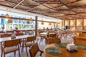 Un amplio comedor interior con varias mesas y sillas, un diseño moderno en el techo y grandes ventanales que ofrecen vistas al exterior.