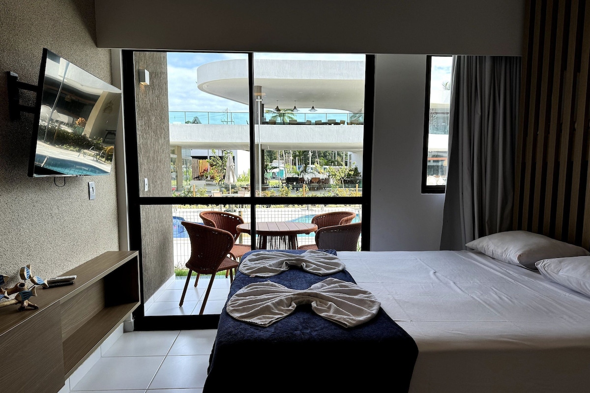 A hotel room with a bed, a TV mounted on the wall, a balcony with chairs, and a view of the outside.
