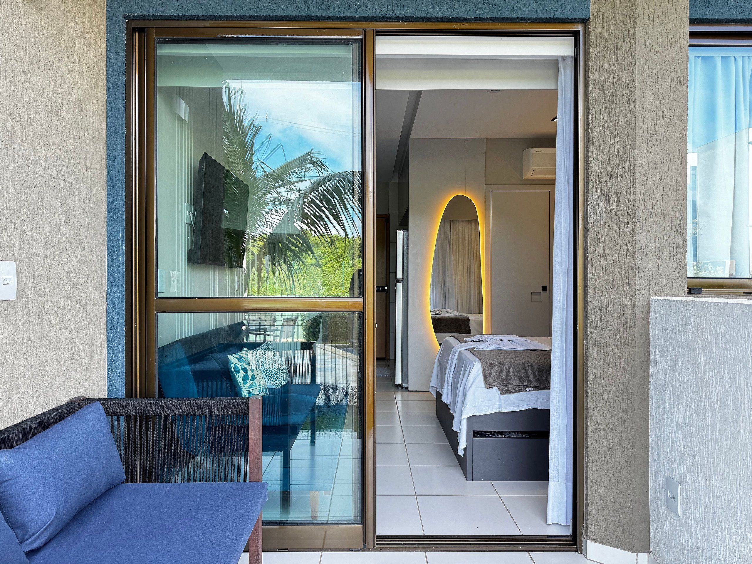 A modern hotel room with a glass door leading to a balcony, a bed with a white headboard, and a small bedside table.
