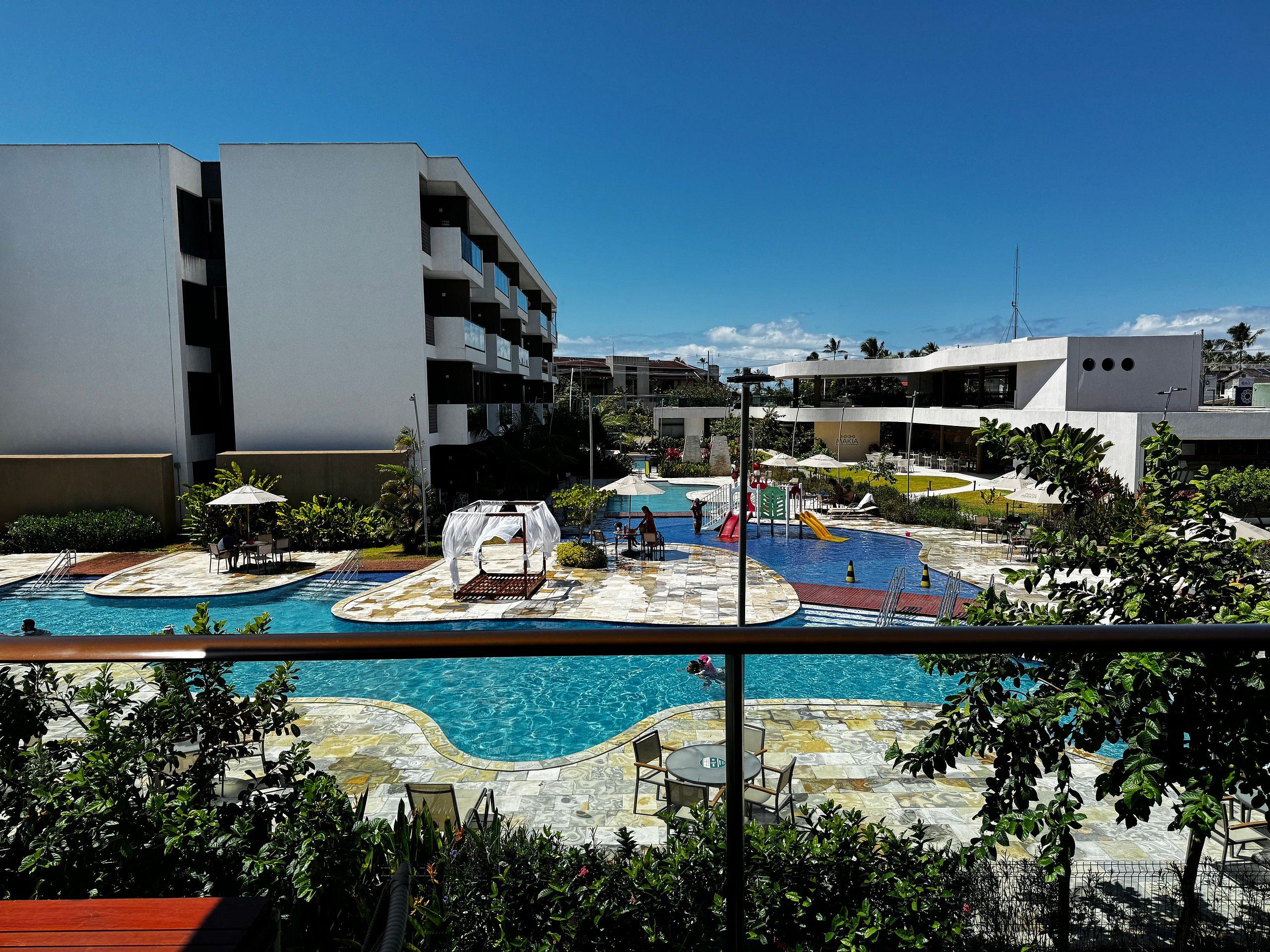 Una piscina con tumbonas y sillones de descanso, rodeada de jardines con palmeras y un edificio moderno al fondo.