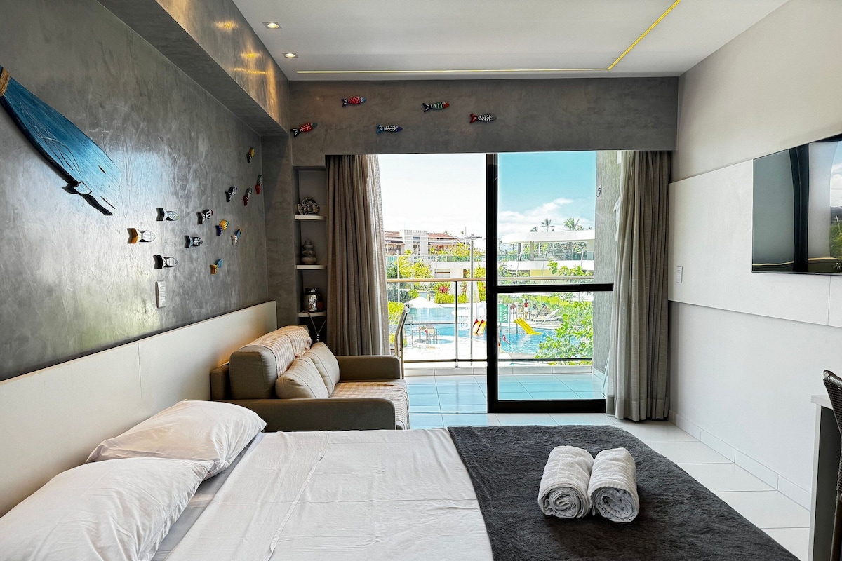 A modern hotel room with a bed, a sofa, a TV, and a balcony with a pool view.