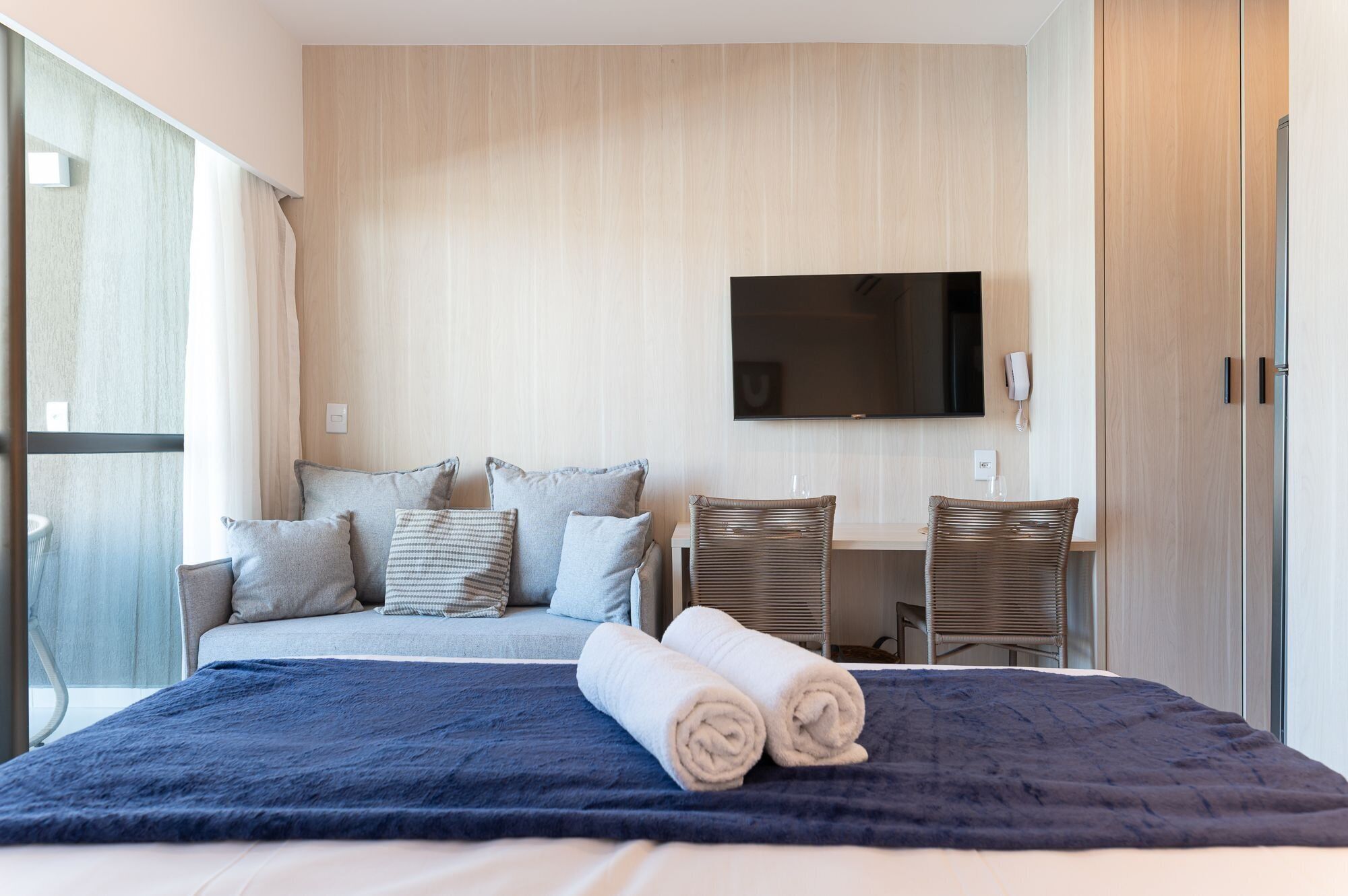 A hotel room with a bed, a flat-screen TV mounted on the wall, a desk with a chair, and a wardrobe.