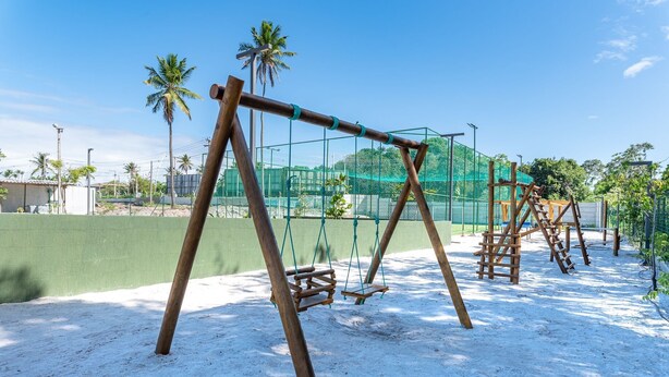 Um parquinho com balanços e estruturas para escalar, cercado por uma grade verde e palmeiras.