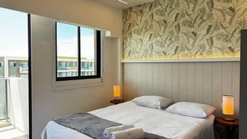 A hotel room with a large bed, two bedside lamps, a window with a view, and a tropical leaf-patterned wall.