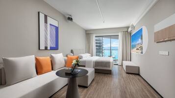 A modern hotel room with a sofa, a bed, a small table with a vase, and a large window offering a view of the sea.