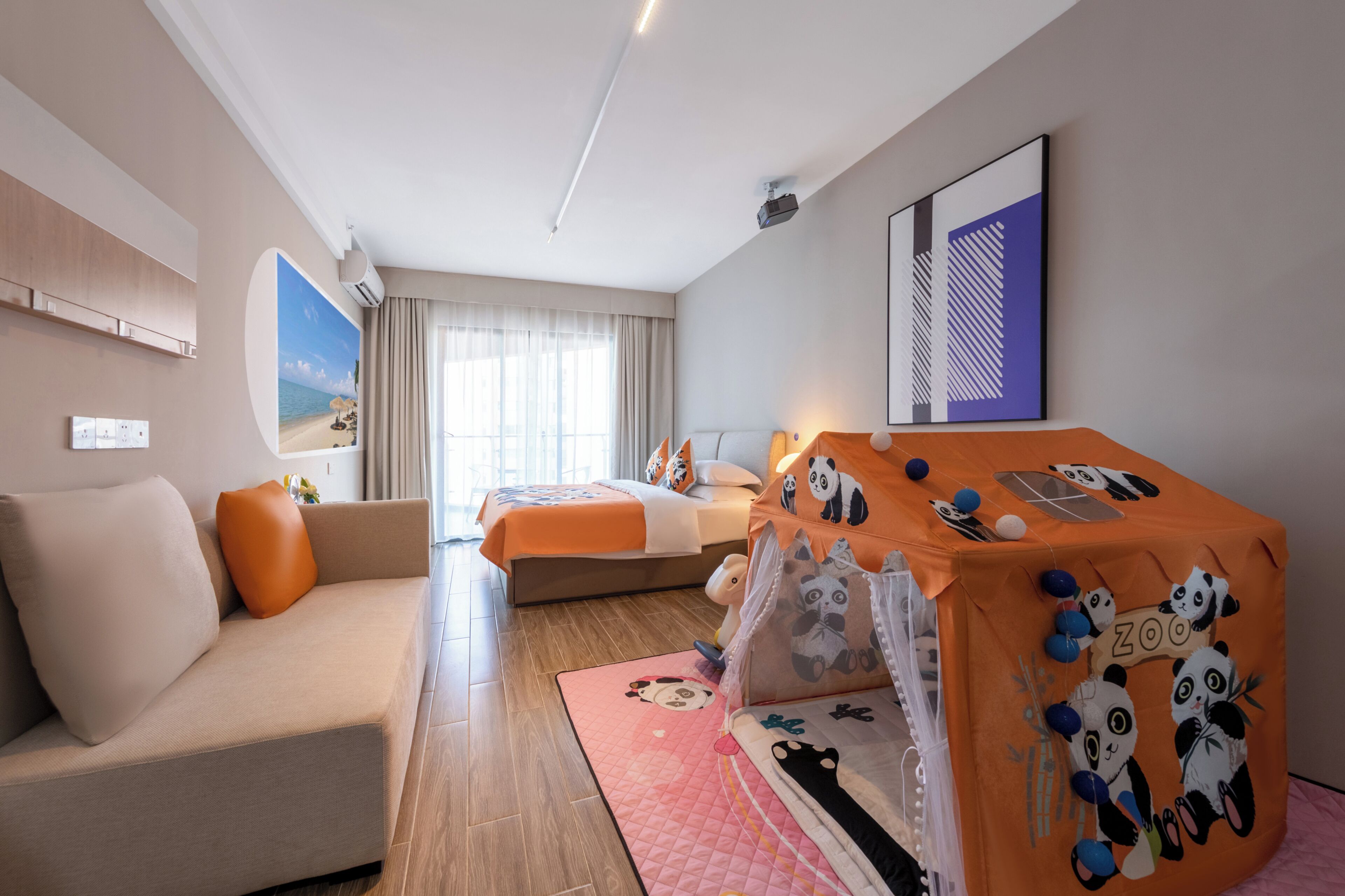 A hotel room with a sofa, a bed with an orange panda-themed bedspread, a small table, and a window with sheer curtains.
