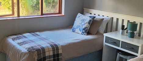 A single bed with a checkered blanket, a pillow with a floral design, and a bedside table with a lamp and remote controls.
