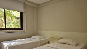 Ein Zimmer mit zwei Betten, einem Fenster mit Blick auf Grünflächen und einem Nachttisch mit Lampe.