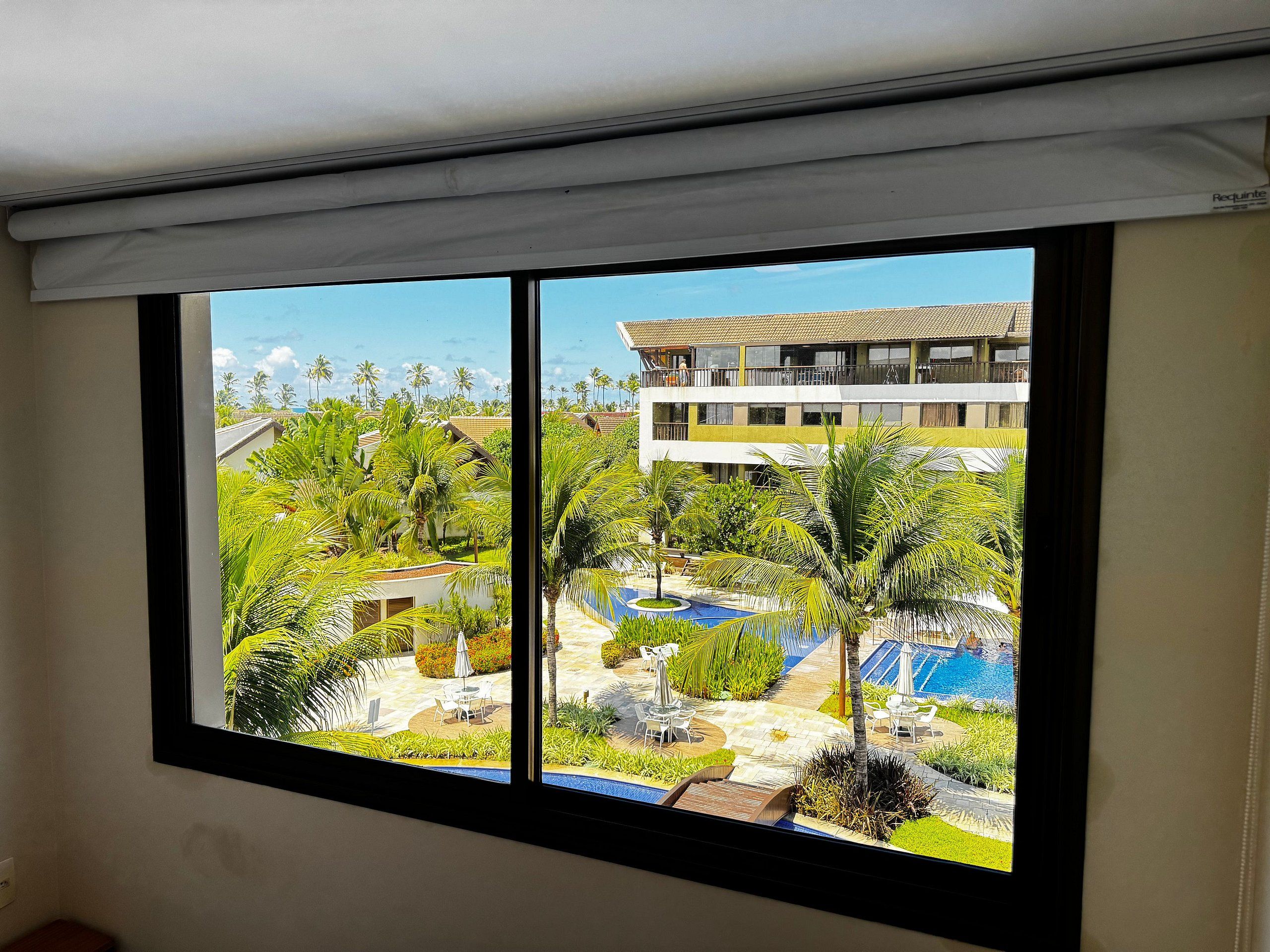 Une chambre d’hôtel avec une grande fenêtre donnant sur un espace piscine agrémenté de palmiers et sur un bâtiment en arrière-plan.