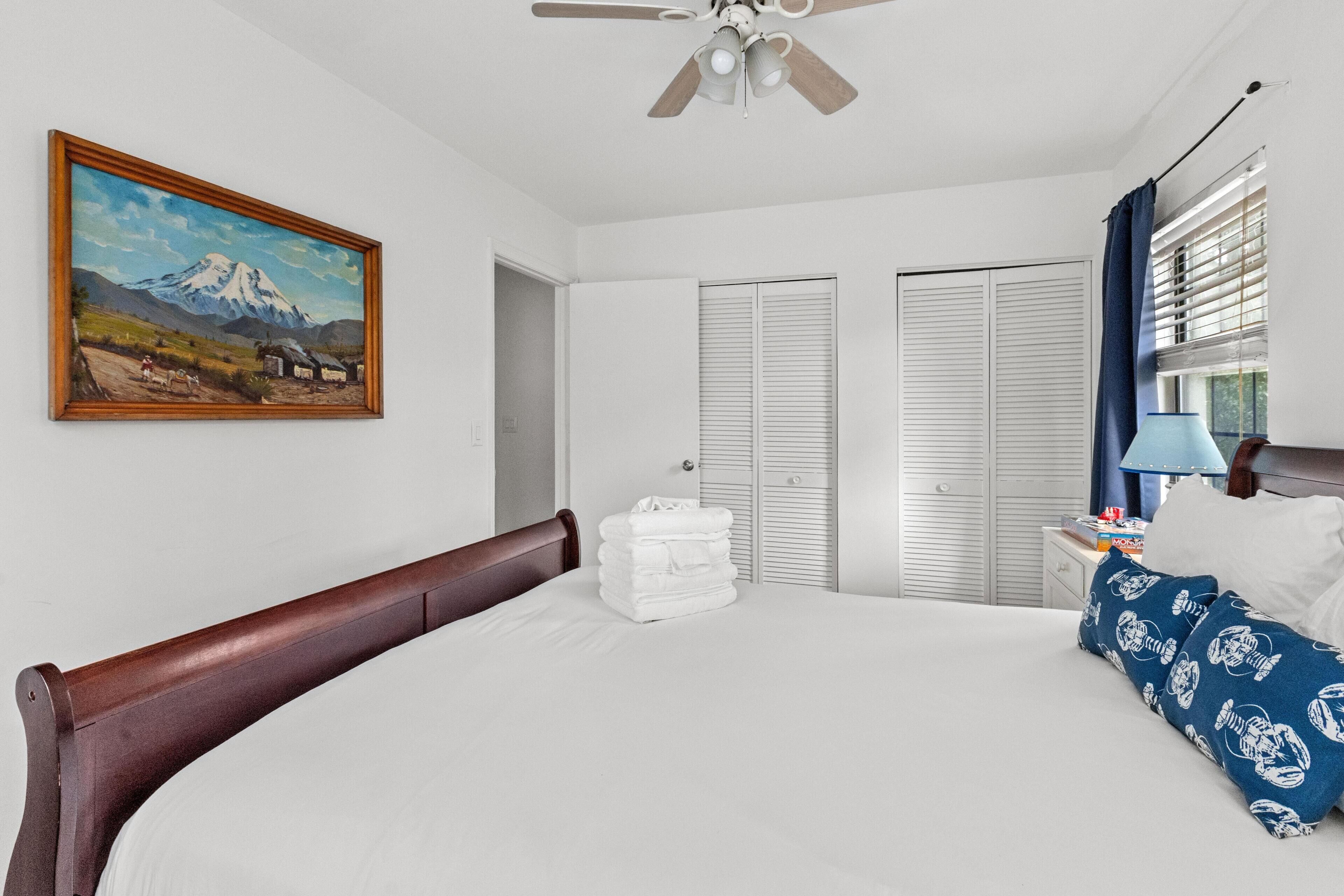 Une chambre à coucher avec un grand lit, un ventilateur de plafond, un tableau au mur et une fenêtre avec des stores.