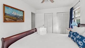 Une chambre à coucher avec un grand lit, un ventilateur de plafond, un tableau au mur et une fenêtre avec des stores.