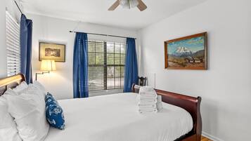 Un dormitorio con cama, ventilador de techo, ventana con persianas, lámpara y un cuadro en la pared.