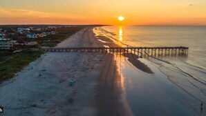 Een strand met een pier die het water in reikt, een zonsondergang op de achtergrond en gebouwen in de verte.