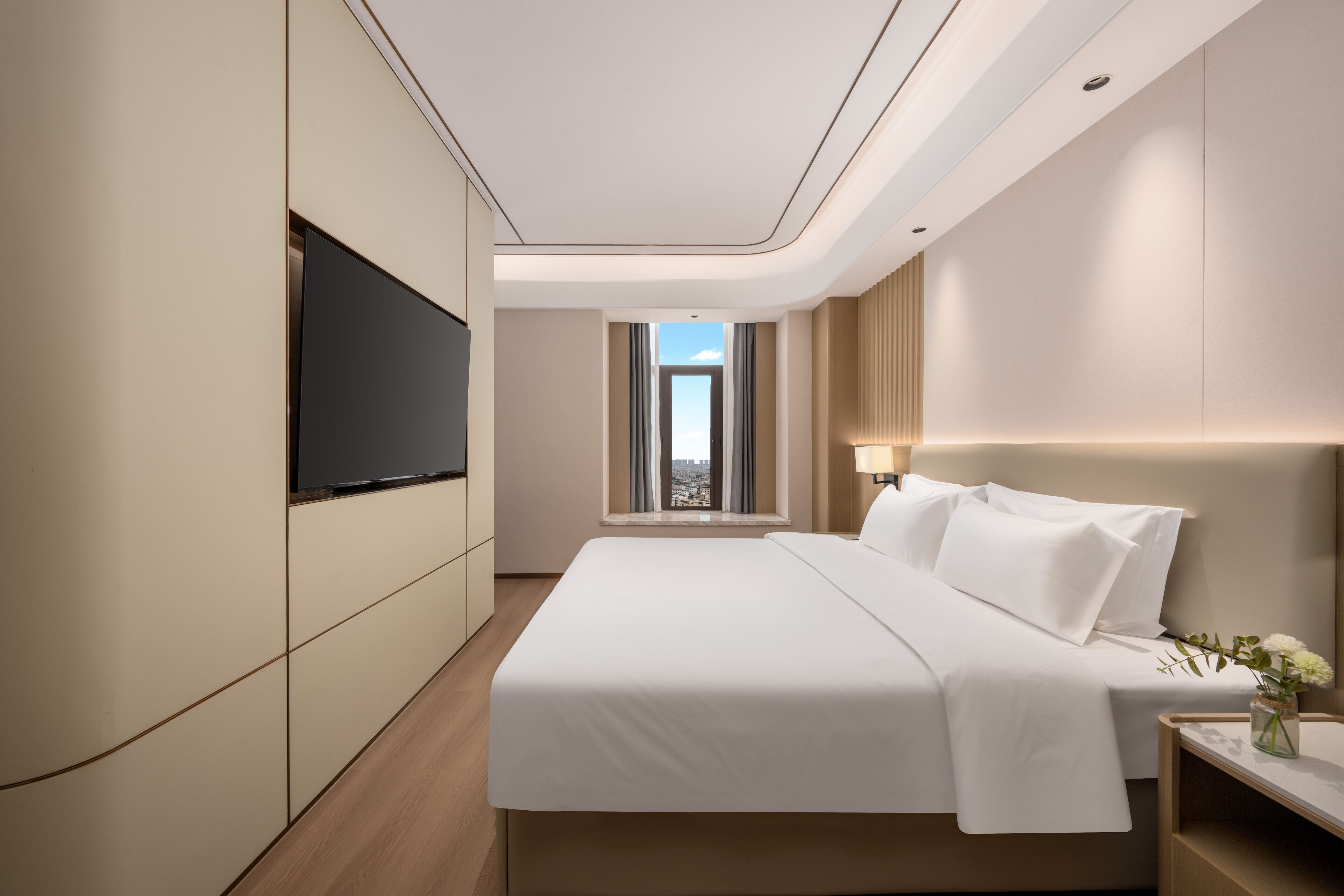 A modern hotel room with a large bed, a flat-screen TV mounted on the wall, a bedside table with a lamp, and a view of the city through the window.