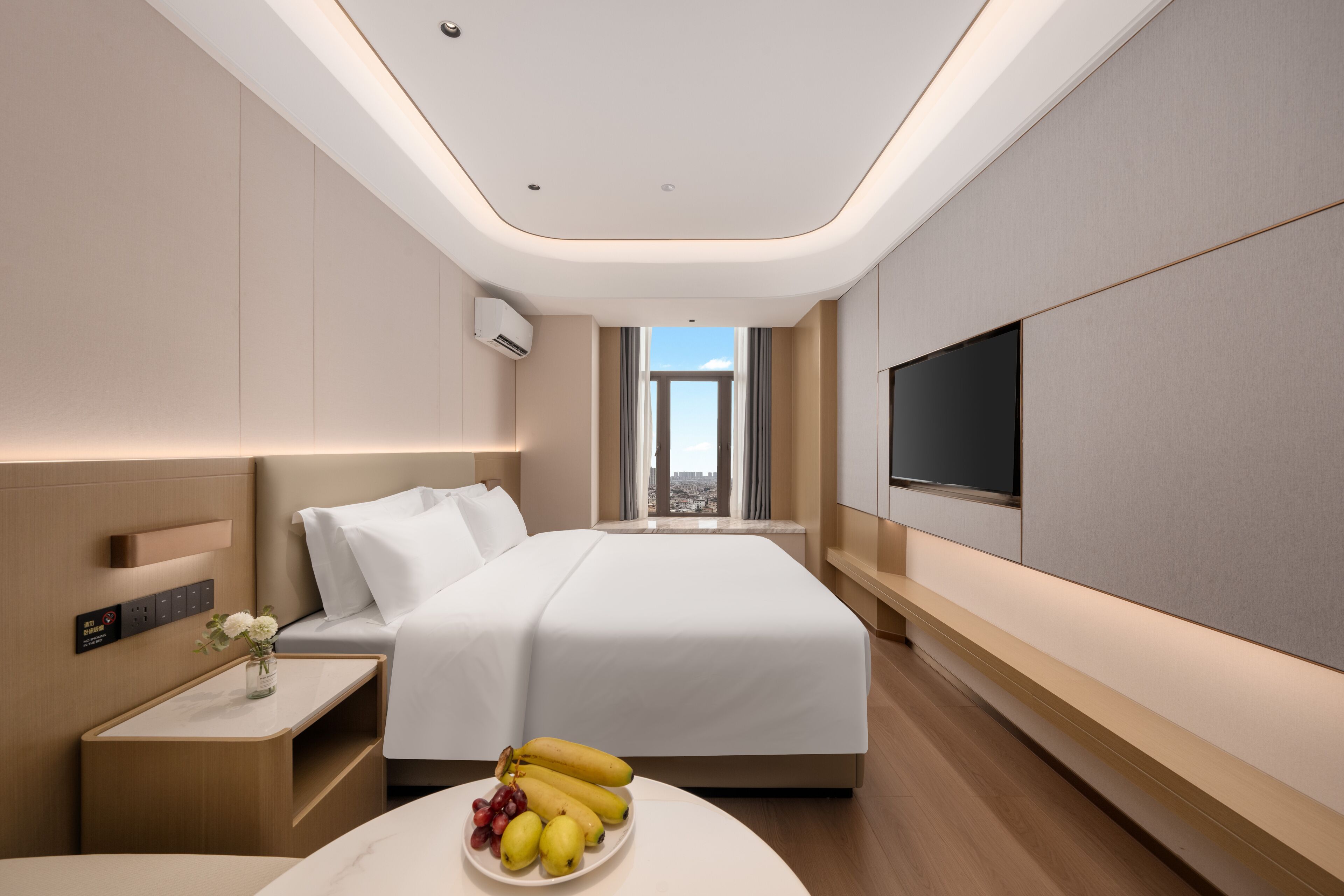 A modern hotel room with a large bed, a flat-screen TV, a bedside table with a vase of flowers, and a view of the city through the window.