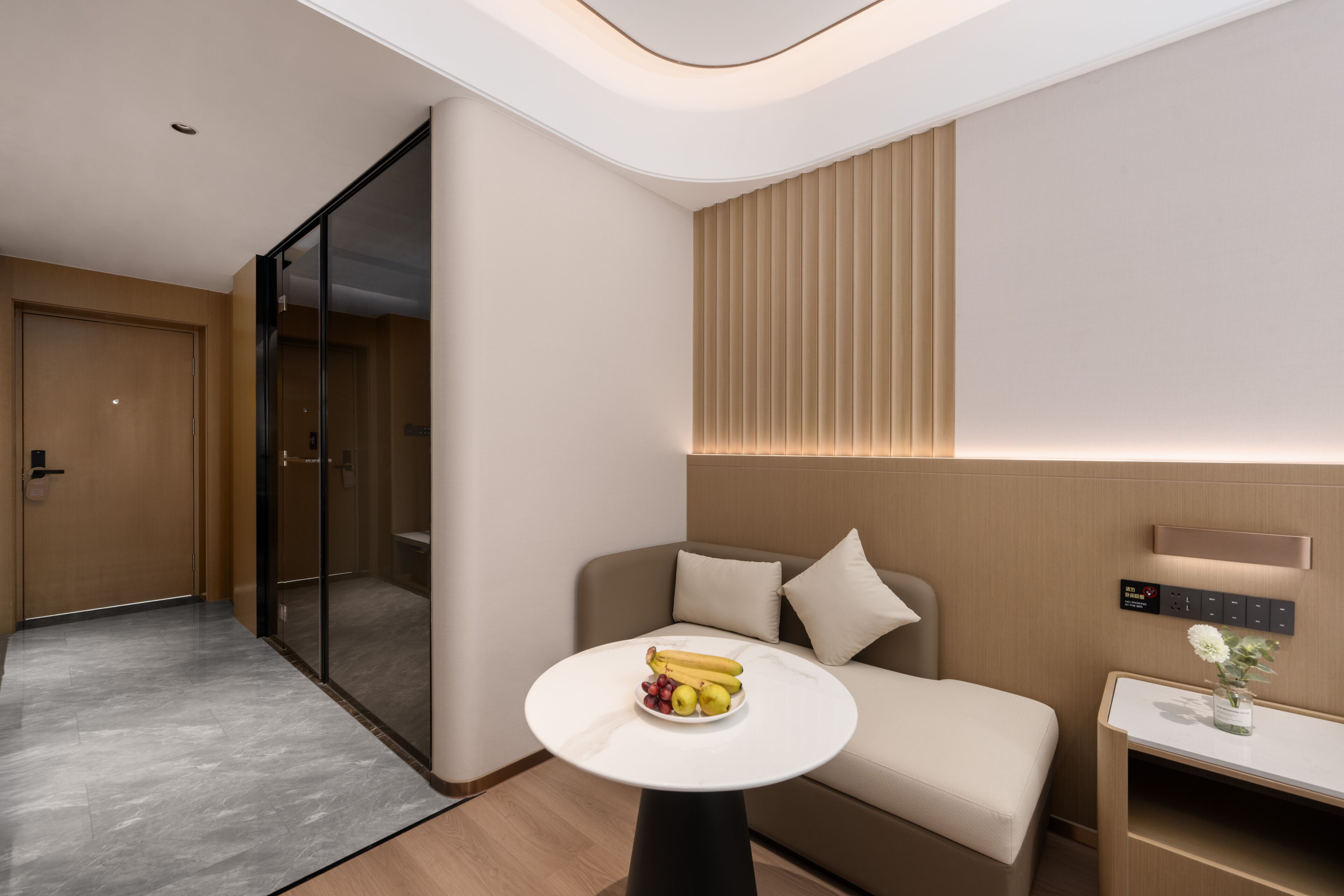 A modern hotel room with a sofa, a small table with fruit, a bedside table with a vase, and a wall-mounted control panel.