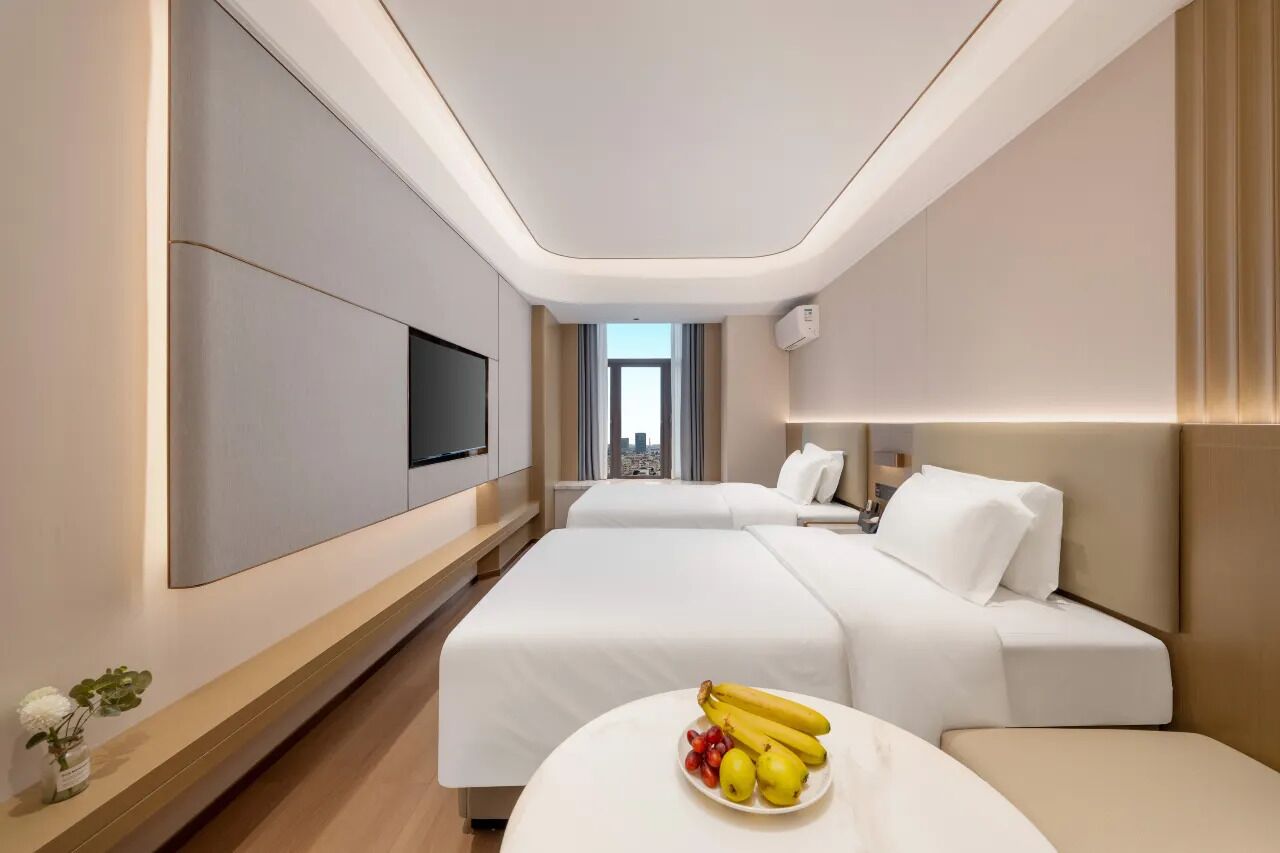 A hotel room with two beds, a flat-screen TV, a city view, and a fruit bowl on the nightstand.