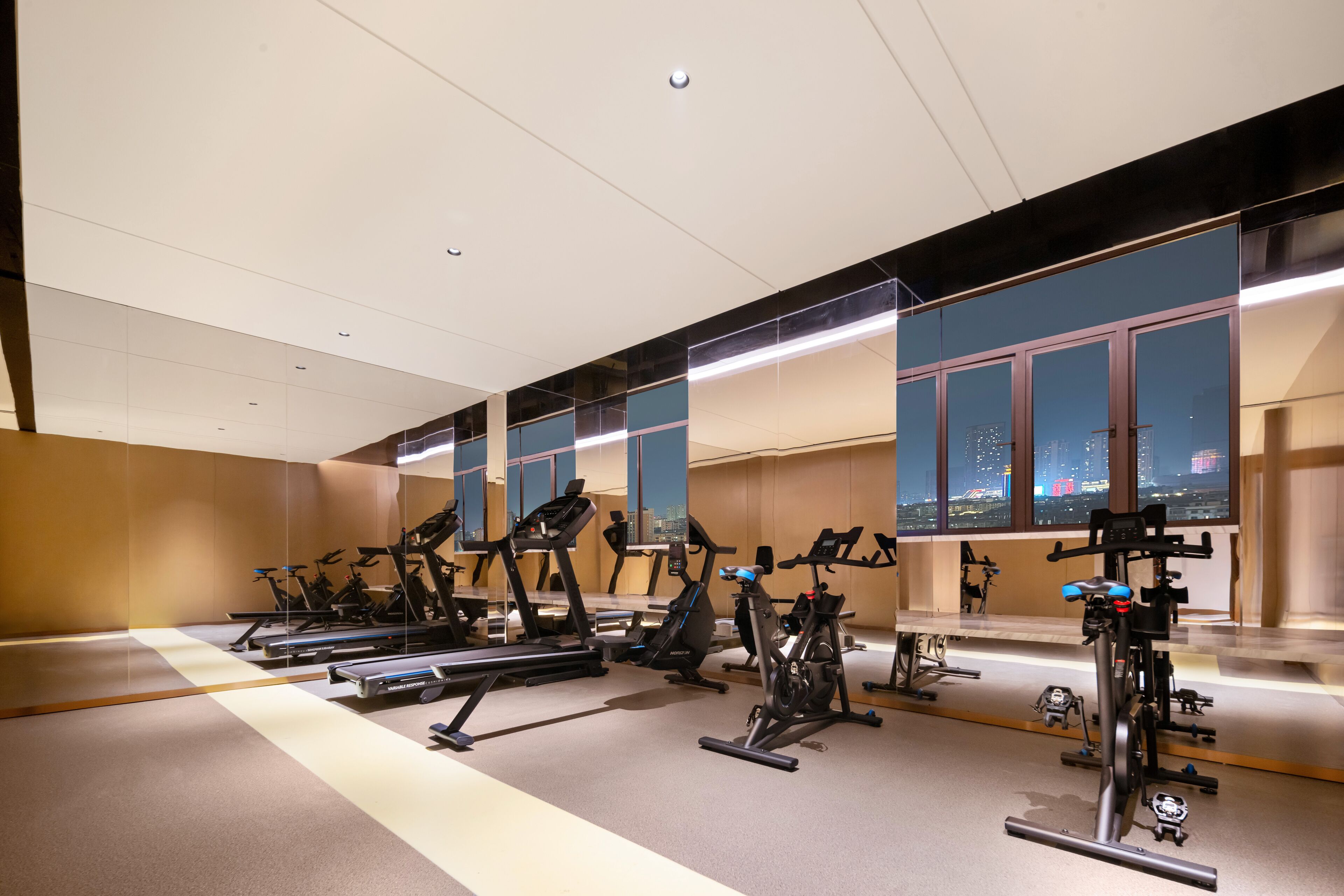 A gym with multiple exercise bikes and treadmills, large windows with a city view, and a high ceiling.