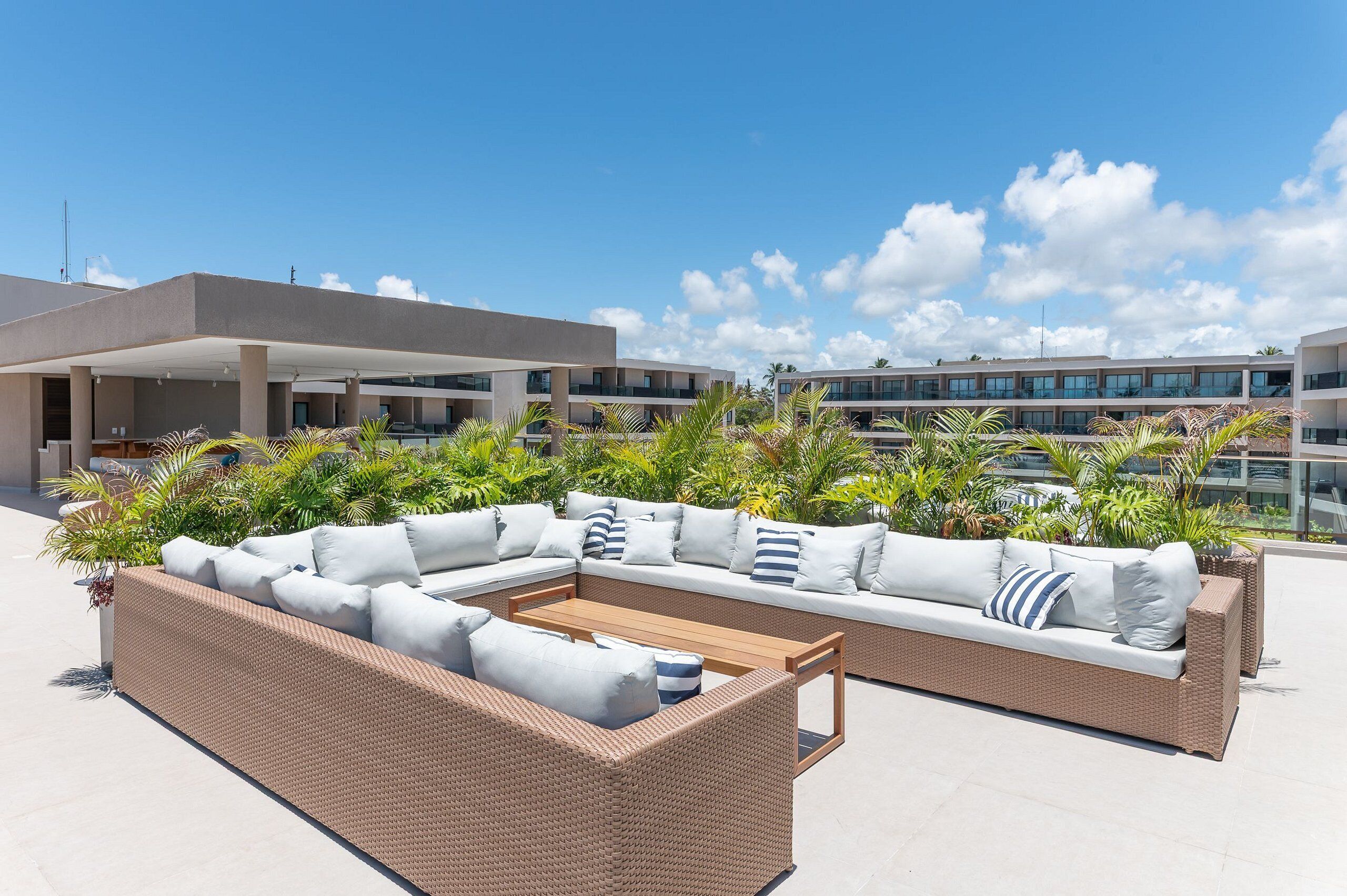 A rooftop terrace with a sectional sofa, a wooden coffee table, and lush tropical plants.