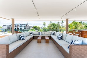 Un espacio de descanso al aire libre con techo, un amplio sofá modular, una mesa de centro de madera y vistas a edificios y vegetación.