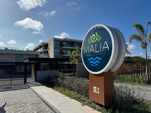Letrero que dice 'MALIA Beach Experience' con un edificio moderno al fondo.