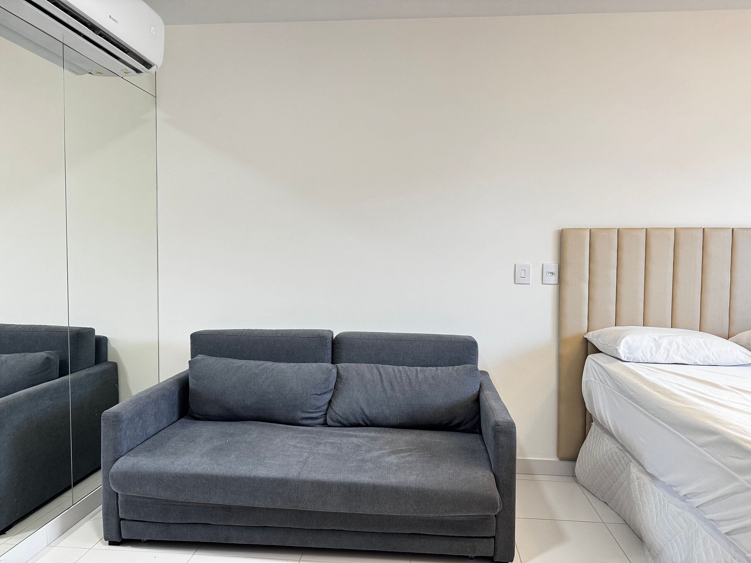 Una habitación moderna con un sofá gris, una cama con ropa de cama blanca y un aire acondicionado de pared.