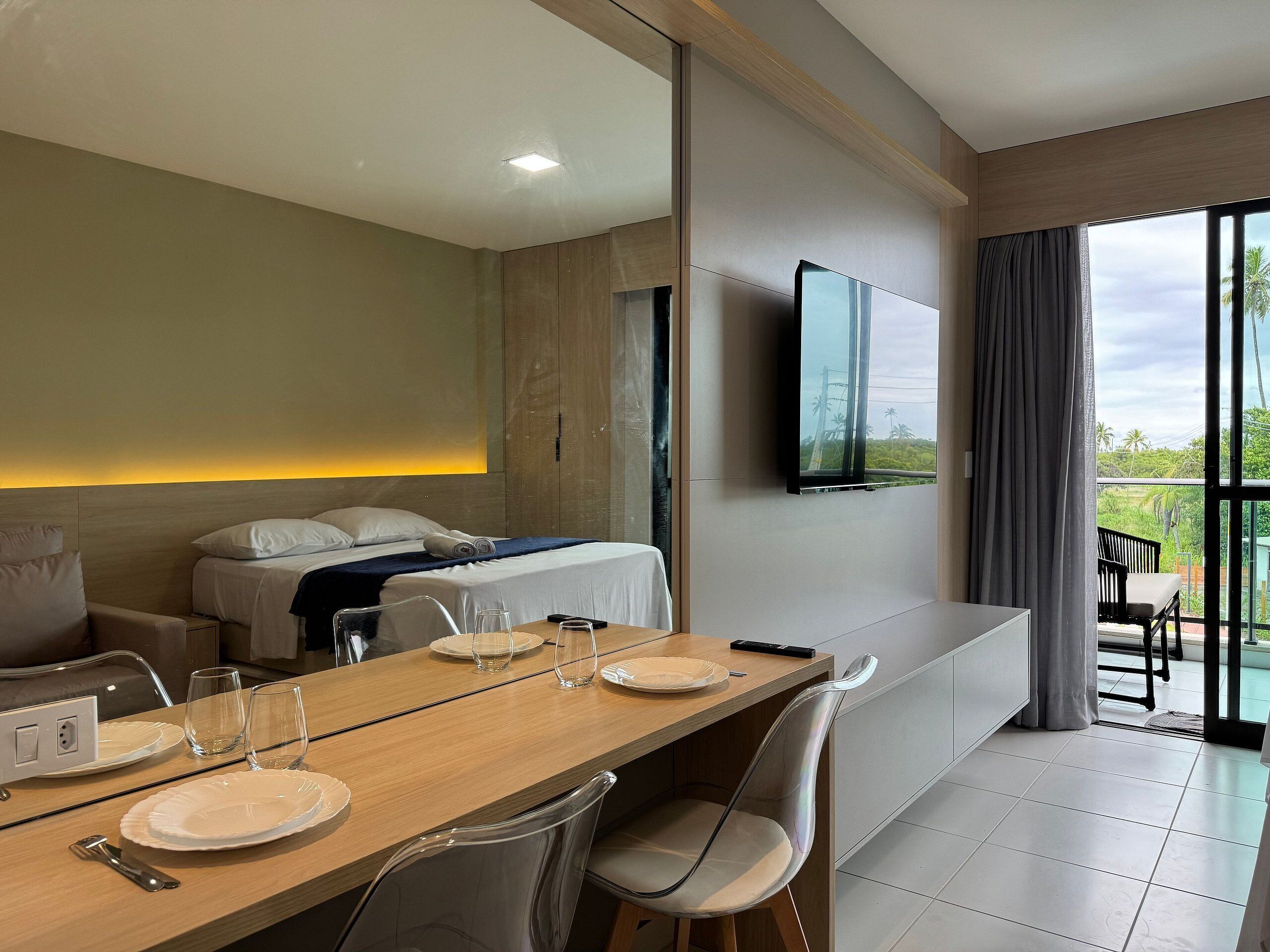 A hotel room with a bed, a dining table set for two, a TV mounted on the wall, and a balcony with outdoor furniture.