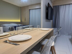 A hotel room with a dining table set for two, a bed with a headboard, a flat-screen TV mounted on the wall, and a window with curtains. - Malia Beach Experience por Brevelar (Ipojuca)