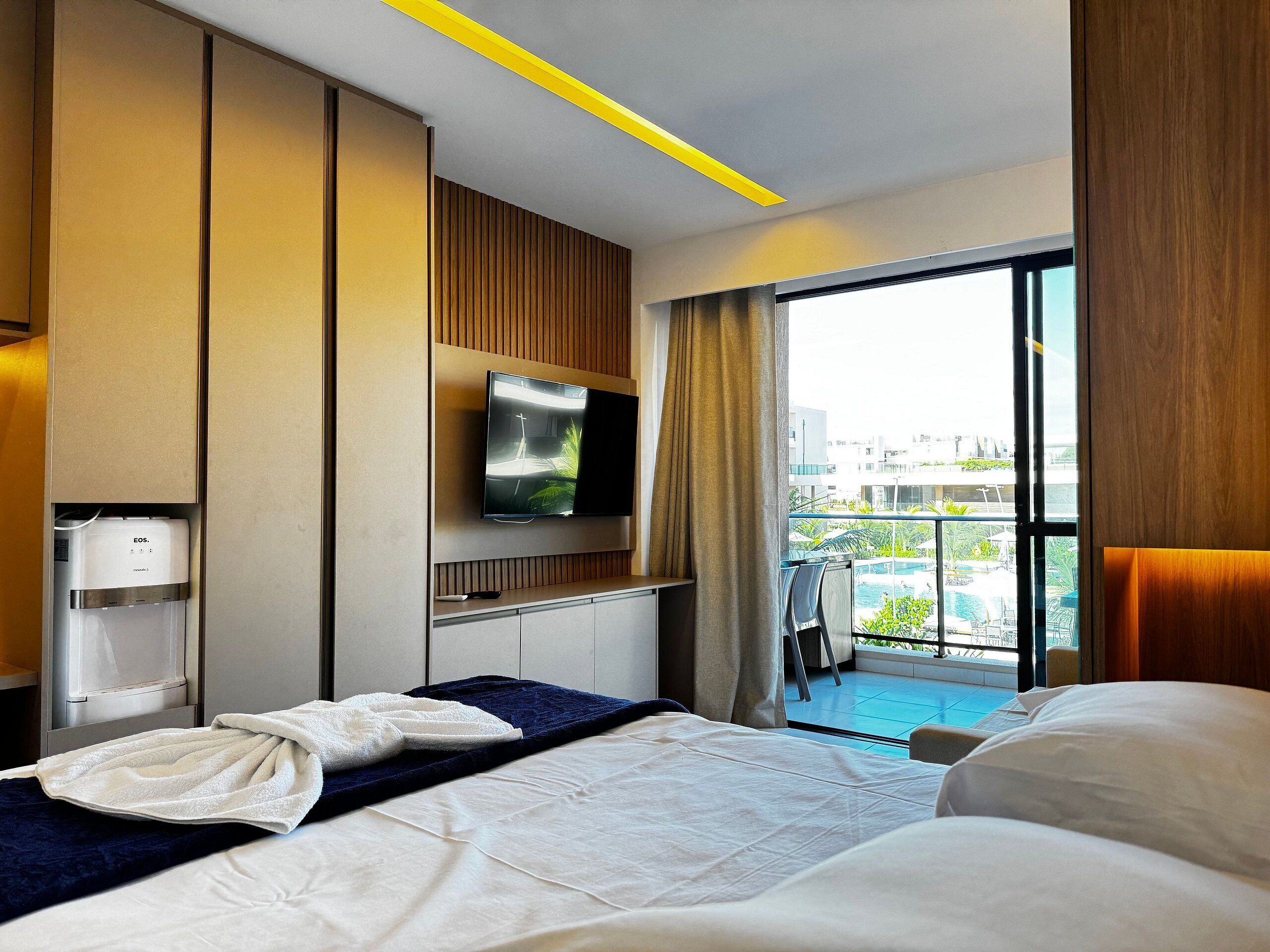 Quarto de hotel com cama, televisão, dispensador de água e vista para uma piscina ao ar livre.