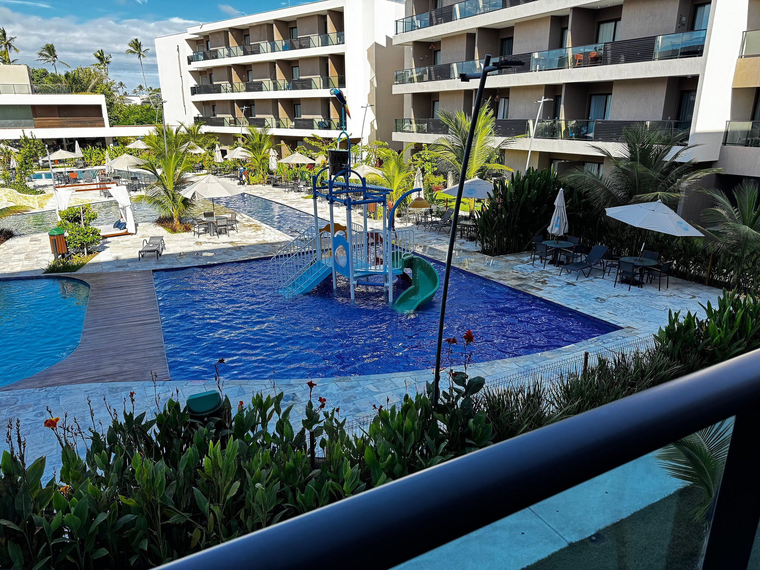 A hotel pool area with a children's play structure, lounge chairs, and a multi-story building in the background.
