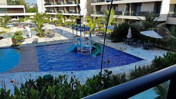 A hotel pool area with a children's play structure, lounge chairs, and a multi-story building in the background.