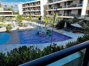 A hotel pool area with a children's play structure, lounge chairs, and a multi-story building in the background.