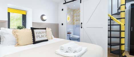 A bedroom with a bed, pillows, a bedside table, a wall-mounted light, a window with a view, and a door leading to a bathroom.