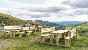 Un área de picnic con bancos de madera y mesas de picnic en una ladera cubierta de césped, con vistas panorámicas de colinas onduladas y un muro de piedra al fondo.