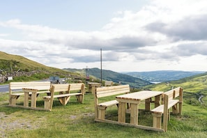 Ein Picknickbereich mit Holzbänken und Picknicktischen auf einer grasbewachsenen Anhöhe, mit schöner Aussicht auf sanfte Hügel und einer Steinmauer im Hintergrund.