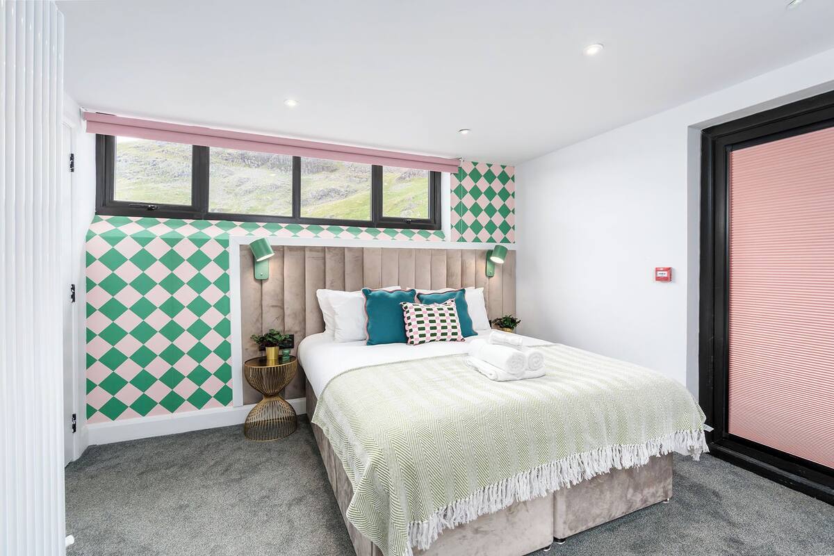 A bedroom with a bed, a window with a view, a wall with a geometric pattern, and a door with a red handle.