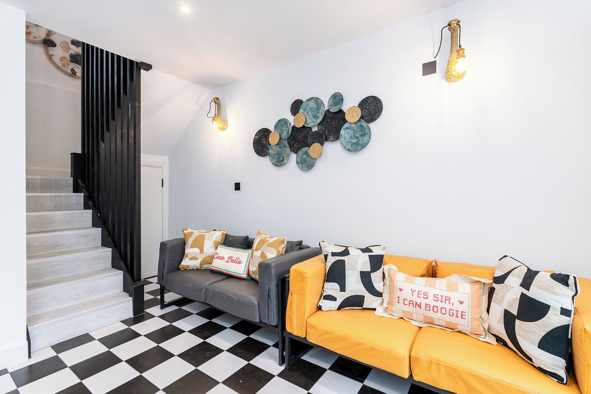 A modern living room with a checkered floor, a grey sofa with yellow cushions, and a black staircase.