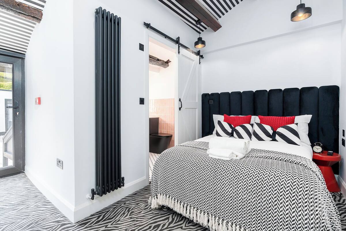 A modern bedroom with a black and white patterned bedspread, a black radiator, and a wooden ceiling beam.