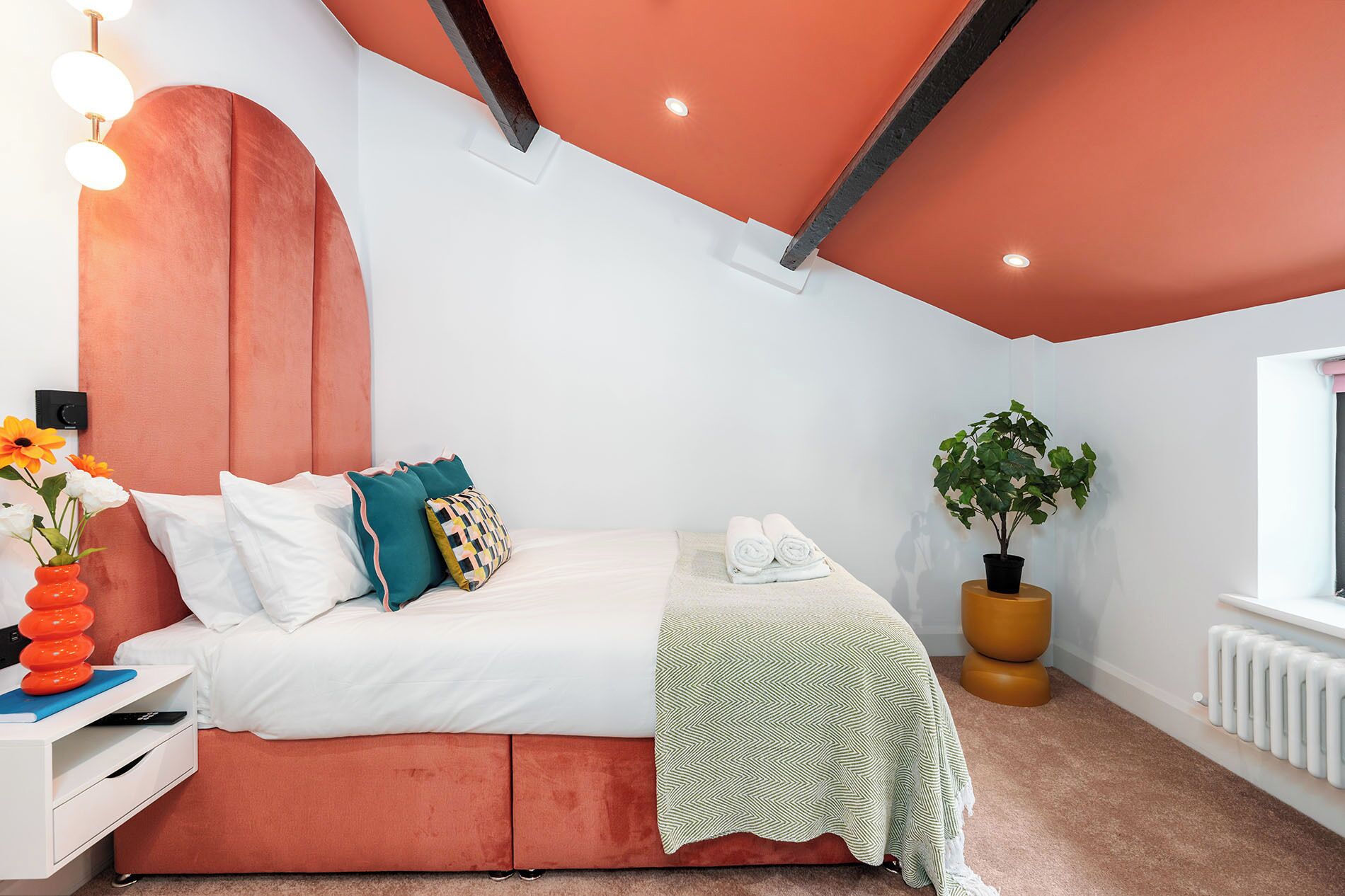 Un dormitorio moderno con una pared de acento rosa, una cama con ropa blanca, almohadas verde azulado y amarillas, un plaid verde estampado, una planta en maceta y una mesita de noche con un florero.