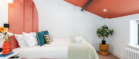 Un dormitorio moderno con una pared de acento rosa, una cama con ropa blanca, almohadas verde azulado y amarillas, un plaid verde estampado, una planta en maceta y una mesita de noche con un florero.