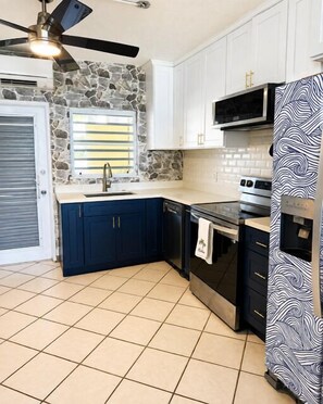 A kitchen with white cabinets, a stone accent wall, a microwave, and a patterned refrigerator. - Welcome to Island Breeze, Your Oceanfront Escape in Beautiful St. Croix (Christiansted)