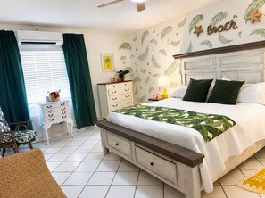 A bedroom with a bed, a nightstand, a chair, a dresser, and a wall with a beach-themed design. - Welcome to Island Breeze, Your Oceanfront Escape in Beautiful St. Croix (Christiansted)