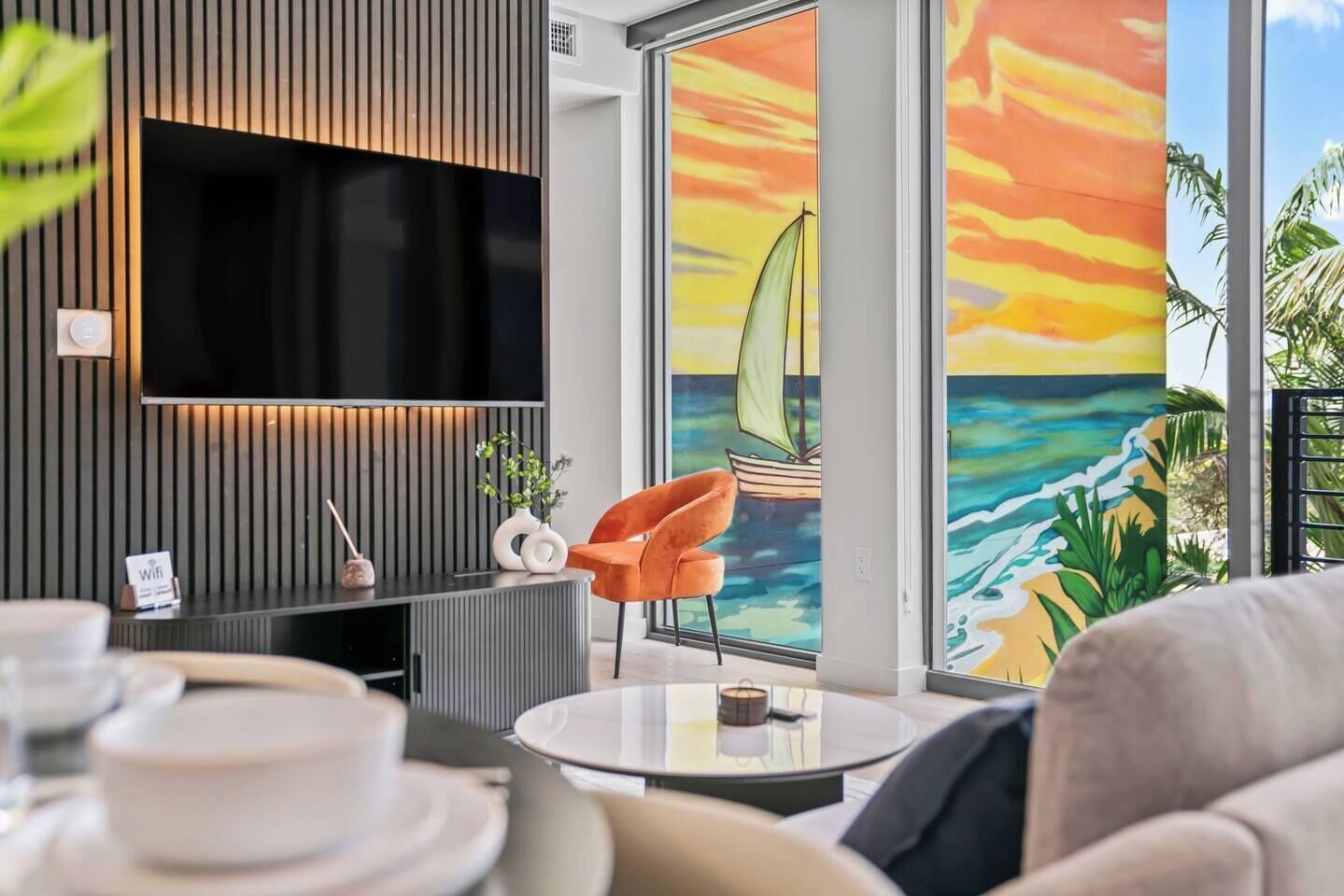 Un salon moderne avec un téléviseur à écran plat, une table basse ronde et une fresque murale aux couleurs vives représentant un coucher de soleil.