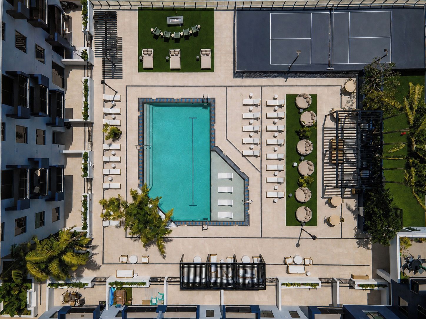 An aerial view of a hotel with a swimming pool, sun loungers, a tennis court, and a landscaped garden.