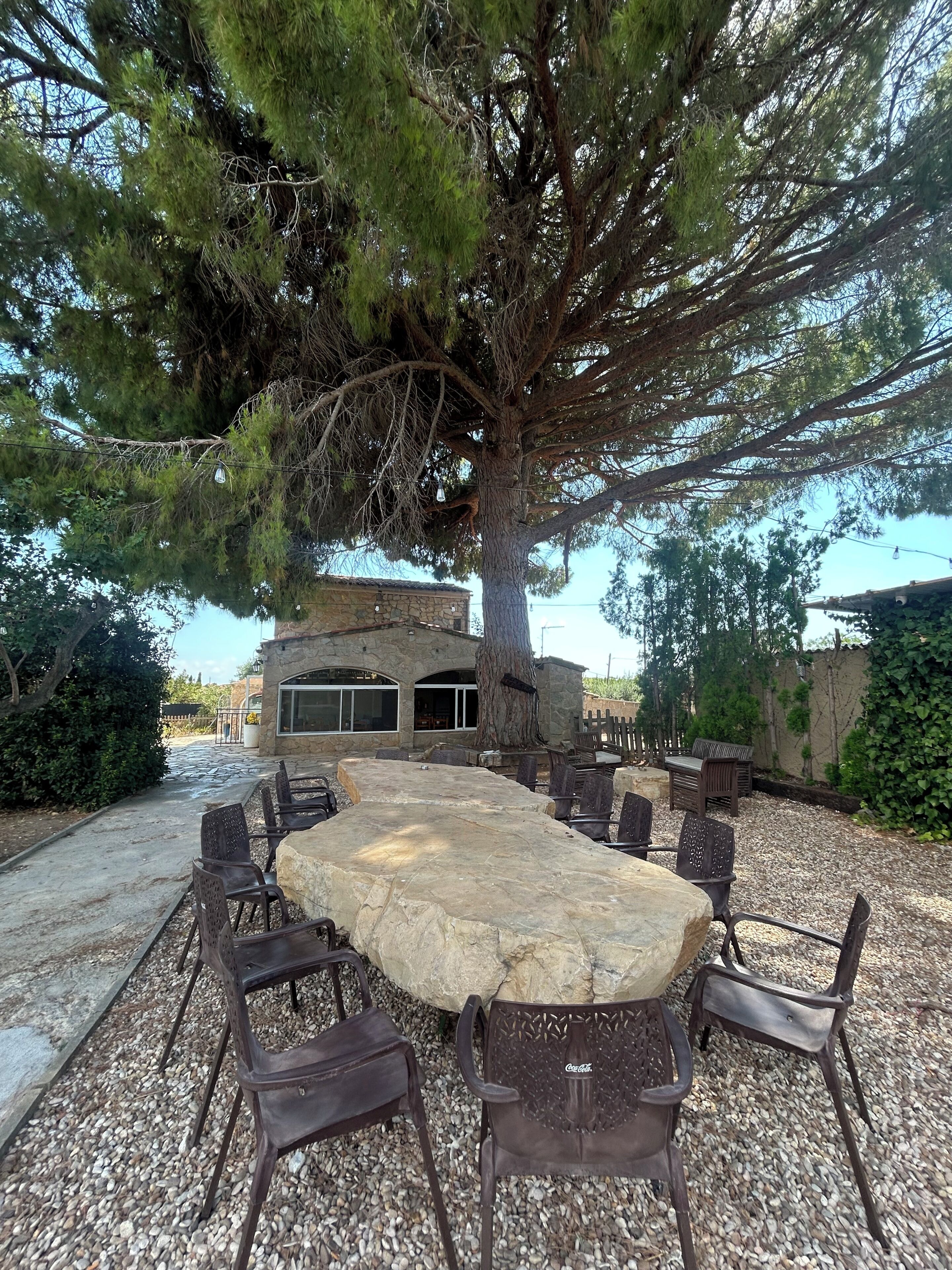 An outdoor patio with a stone table and metal chairs, surrounded by a gravel area and a large tree.