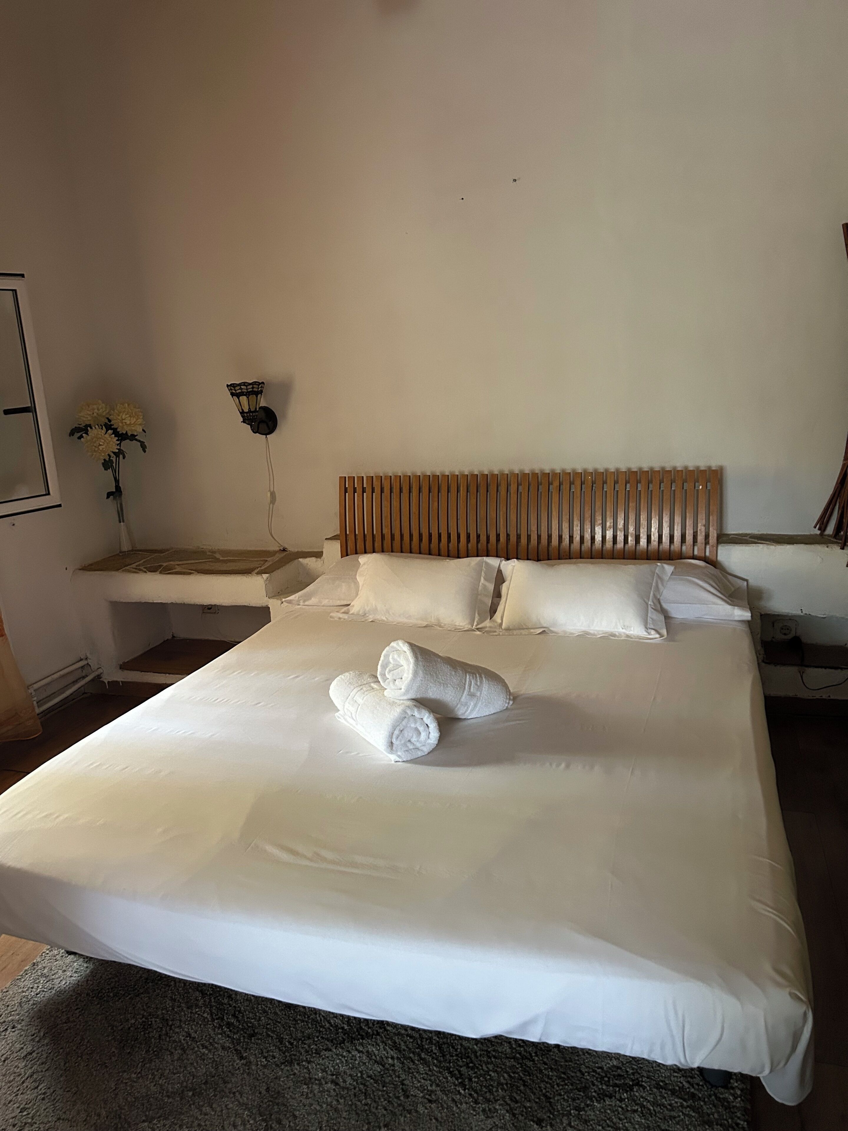 Une chambre d’hôtel avec un grand lit, deux serviettes roulées, une tête de lit en bois, une table de chevet ornée d’un vase de fleurs et une lampe fixée au mur.