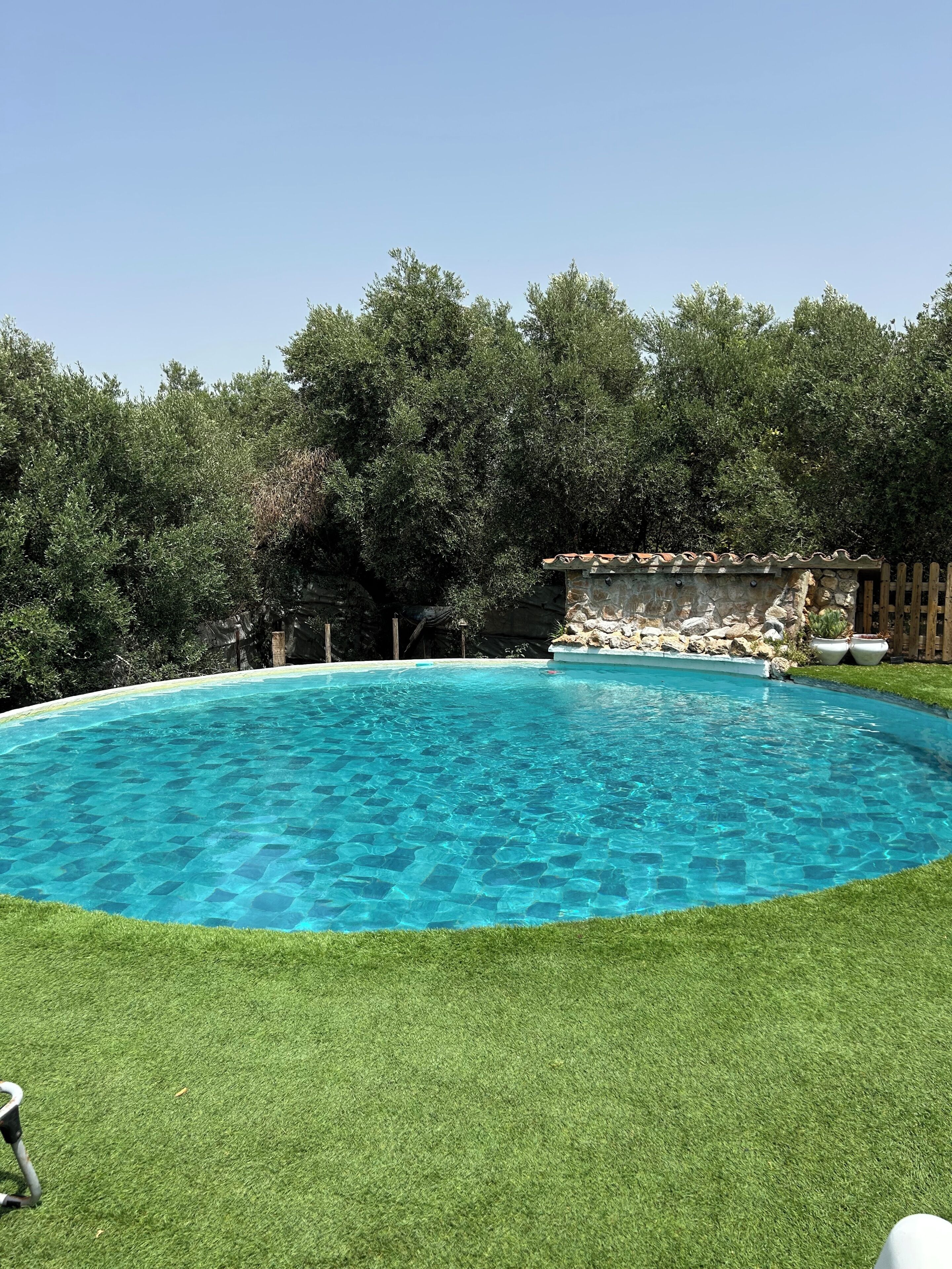 Uma piscina azul límpida rodeada por um relvado bem cuidado, com um muro de pedra e árvores ao fundo.